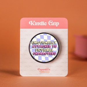 Peut inclure: Une poignée de téléphone à damiers noir et blanc avec le texte "Emotionally Attached to Fictional Characters" en lettres roses et jaunes. La poignée est sur une carte blanche avec le texte "Kindle Grip" en lettres roses et "Paperback Addiction" en lettres noires.