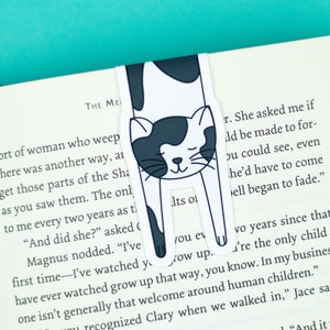 May include: A black and white cat bookmark with a sleeping cat illustration. The bookmark is placed in a book with the text "THE ME" and "sort of woman who weep" visible. The text on the bookmark reads "Its ol" and "you ev".