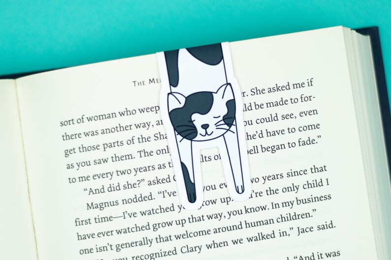 May include: A black and white cat bookmark with a sleeping cat illustration. The bookmark is placed in a book with the text "THE ME" and "sort of woman who weep" visible. The text on the bookmark reads "Its ol" and "you ev".