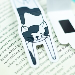 May include: Two black and white cat-shaped magnetic bookmarks. The cat is curled up with its eyes closed. The bookmarks are on a book.