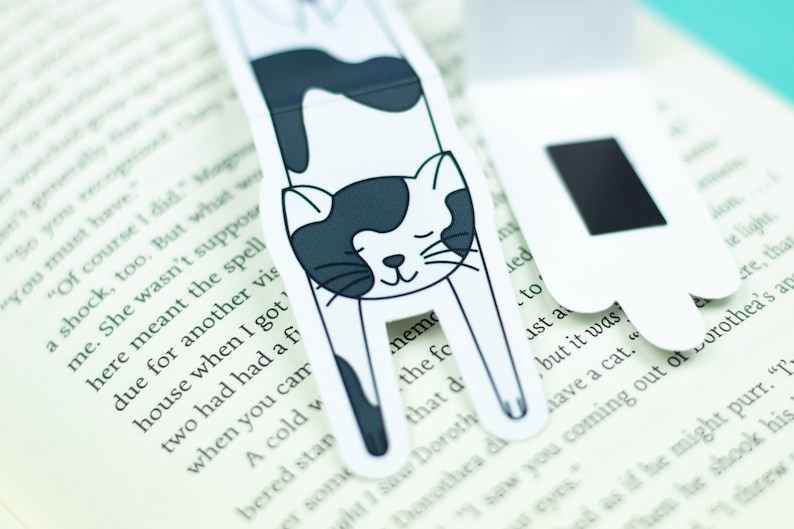 May include: Two black and white cat-shaped magnetic bookmarks. The cat is curled up with its eyes closed. The bookmarks are on a book.