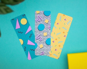 Pode incluir: Três marcadores de livros coloridos com padrões geométricos. Os marcadores têm um fundo azul, amarelo e branco com formas geométricas rosa, amarelas e azuis.