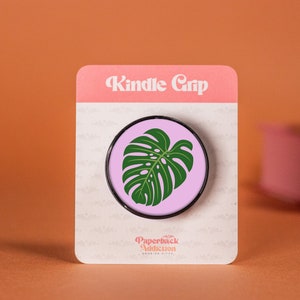 Puede incluir: Un agarre para teléfono de color rosa y negro con un diseño de hoja de monstera verde. El agarre está empaquetado en una tarjeta blanca con el texto "Kindle Grip" y "Paperback Addiction".