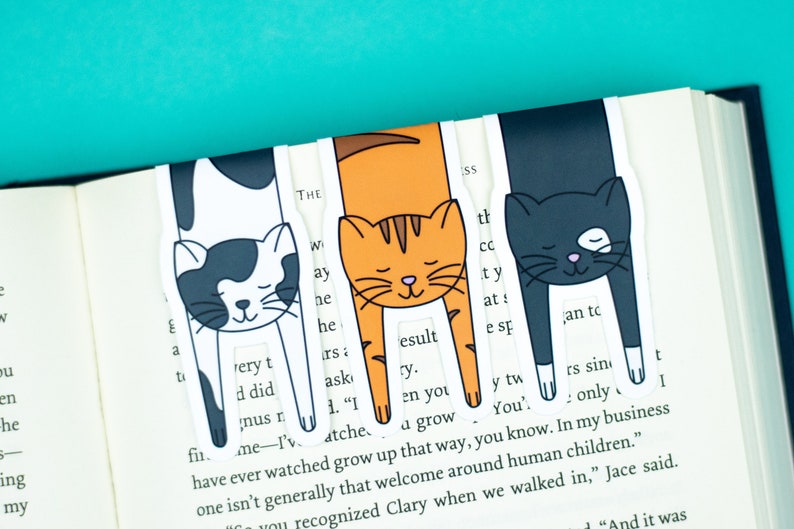 May include: Three cat-shaped bookmarks with black and white, orange and white, and black fur designs. The bookmarks are placed on a book with text.