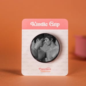 May include: A Kindle Grip featuring a monochrome image of two men in an embrace. The grip is mounted on a pink and white card, with the text "Kindle Grip" and "Paperback Addiction". The background is orange.