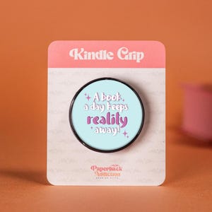 Puede incluir: Un Kindle Grip con un diseño circular azul claro con el texto "A book a day keeps reality away!" en blanco y morado. El agarre está en una tarjeta rosa y blanca con las palabras "Kindle Grip".