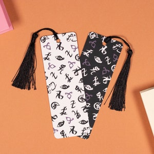 May include: Two black and white bookmarks with a tassel. The bookmarks are covered in a pattern of runes and symbols. One bookmark is white with black runes and the other is black with white runes.