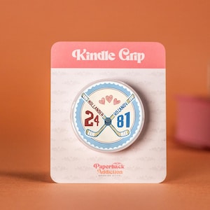 May include: A circular Kindle Grip phone accessory on a rectangular backing card. The design features two crossed hockey sticks, the names Hollander and Rozanov, the numbers 24 and 81, and three small hearts. The text Kindle Grip and Paperback Addiction are printed on the card.