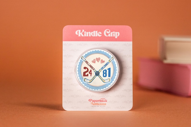 May include: A Kindle Grip with a hockey theme. The round grip features crossed hockey sticks, the numbers 24 and 81, and the names Hollander and Rozanov. The grip is attached to a pink and white backing with the words "Kindle Grip".