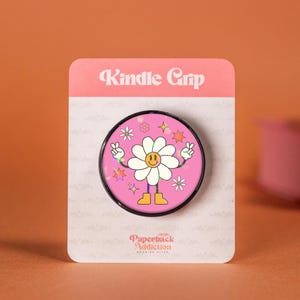 Puede incluir: Un agarre para teléfono de color rosa y negro con un diseño de margarita sonriente. La margarita tiene signos de paz para las manos y está rodeada de purpurina y estrellas. El texto "Kindle Grip" está en la parte superior del embalaje y "Paperback Addiction" en la parte inferior.