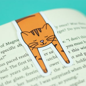 May include: Orange and brown cat bookmark with a white background. The cat is curled up with its head resting on the page. The bookmark is designed to hold a page in a book.