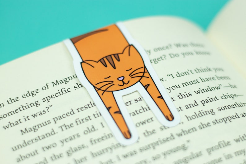 May include: Orange and brown cat bookmark with a white background. The cat is curled up with its head resting on the page. The bookmark is designed to hold a page in a book.