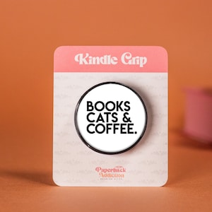 Könnte beinhalten: Ein runder Handy-Griff in Weiß und Schwarz mit dem Text "BOOKS CATS & COFFEE." auf weißem Hintergrund. Der Griff befindet sich auf einer weißen Karte mit dem Text "Kindle Grip" und "Paperback Addiction" in Rosa.