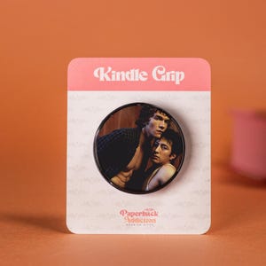 May include: A Kindle Grip featuring a circular image of two men embracing. The grip is on a pink and white card with the text "Kindle Grip" and "Paperback Addiction". The background is orange.