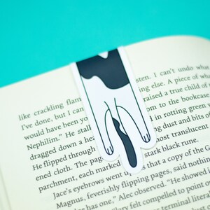 May include: A black and white cat bookmark with a simple design. The bookmark is placed in a book with text about a Nephilim.