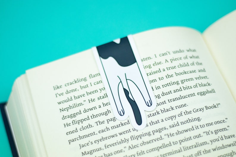 May include: A black and white cat bookmark with a simple design. The bookmark is placed in a book with text about a Nephilim.