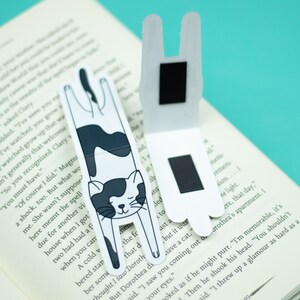 May include: Two black and white cat-shaped magnetic bookmarks. The bookmarks are folded in half and have a black magnet on the back. The cat is upside down with its paws in the air.