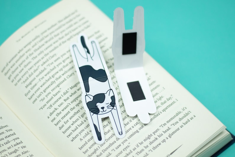 May include: Two black and white cat-shaped magnetic bookmarks. The bookmarks are folded in half and have a black magnet on the back. The cat is upside down with its paws in the air.
