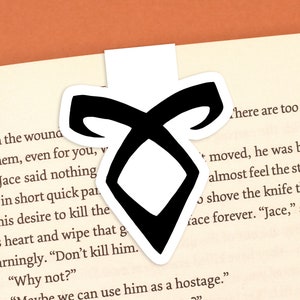 May include: A black and white bookmark featuring the symbol of the Mortal Instruments series, a rune with a double-sided arrow shape.