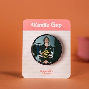 Könnte beinhalten: Ein Kindle Grip mit einem kreisförmigen Bild einer Person in einem schwarzen und goldenen Hockey-Trikot mit dem Logo der Boston Bruins. Der Grip befindet sich auf einer rosa und weißen Karte mit "Kindle Grip" und "Paperback Addiction".