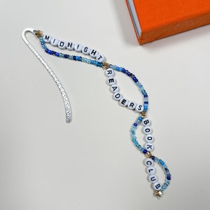May include: A beaded bookmark with the words "MIDNIGHT READERS BOOK CLUB" in white letter beads. The beads are strung on a silver metal bookmark with a decorative design. The beads are in shades of blue and gold.