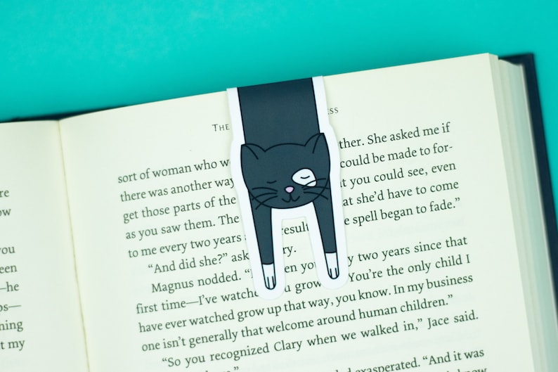 May include: A black cat bookmark with white paws and a white patch on its eye. The bookmark is shaped like a cat hanging upside down with its paws sticking out. The bookmark is on top of an open book with text about a woman and a spell.