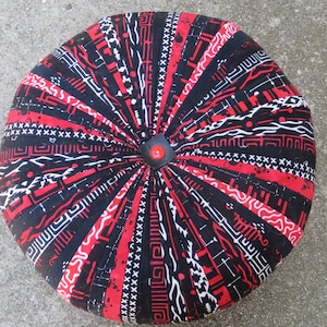 May include: Round, patterned ottoman with a black, red, and white geometric design. The fabric is gathered in a radial pattern, and a small, square, black button with a red center is in the center.