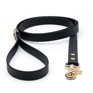 May include: A black dog leash made of a smooth, matte material featuring a gold-toned metal swivel snap hook and a matching gold-toned D-ring near the handle. The leash is coiled in a loop, showcasing the flat, flexible strap and gold rivets.