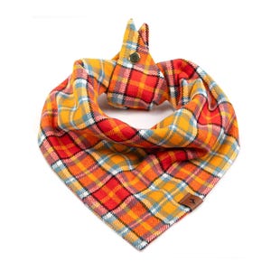 May include: A plaid dog bandana in shades of orange, red, yellow, and blue. The fabric is folded and secured with a snap closure. A small brown leather tag is attached to the bandana.