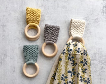 May include: Four wooden rings with attached textured fabric loops in yellow, gray, and blue-green, plus one cream-colored loop holding a white kitchen towel featuring a repeating pattern of blue flowers and green vines on a light background.
