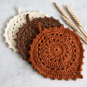 May include: Three crocheted leaf-shaped trivets in cream, brown, and rust-orange. Each trivet has a looped handle and a scalloped edge. The trivets are arranged on a light gray surface, with wheat stalks in the background.
