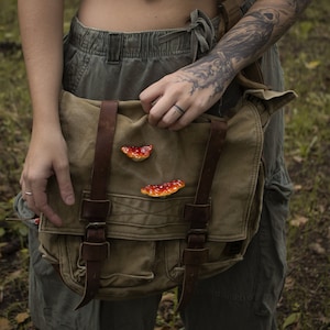 Peut inclure: Une personne portant un pantalon cargo vert et une chemise verte avec un sac messager en toile marron à lanières en cuir. Le sac a trois patchs en forme de champignon rouge et orange attachés à lui.