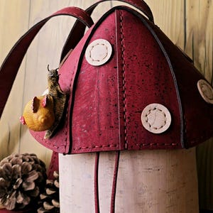 May include: A whimsical mushroom-shaped backpack with a burgundy top and a cream-colored base. The burgundy top features three circular accents and is adorned with a snail figurine. The bag has burgundy straps and drawstrings.