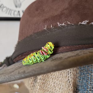 Op de afbeelding: Een bruine vilten fedora hoed met een donkerbruine lintband, voorzien van een klein, gedetailleerd rupsfiguurtje aan de zijkant. De rups is fel limoengroen met zwarte strepen, oranje stippen en kleine witte en rode markeringen op de rug.