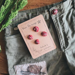 Peut inclure: Boutons rouges à pois blancs en forme de champignons, présentés sur une carte avec l'inscription "Wear the magic". Les boutons sont disposés près d'un short vert olive. Le short comporte un patch avec une illustration d'escargot.