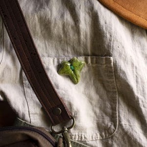 Peut inclure: Une épingle en forme de feuille verte avec un bijou vert au centre, épinglée sur une chemise en lin beige clair avec une sangle en cuir marron et un sac en toile marron.