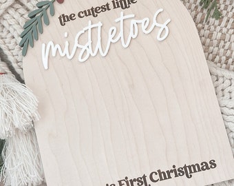 May include: A wooden Christmas sign with the text "the cutest little mistletoes" in white script and "Fallon's First Christmas 2023" in brown. The sign is decorated with green leaves and red berries.