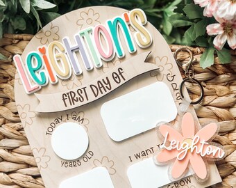 May include: A wooden first and last day of school sign with a pink flower and the name Leighton. The sign has spaces to fill in the child's age, grade, and what they want to be when they grow up.