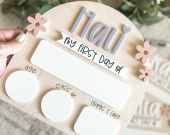 May include: A wooden sign with a light gray arch and the name "Hali" in light gray letters. The sign says "My first day of" and has spaces for the child's age, class, and things they love. The sign is decorated with pink flowers.