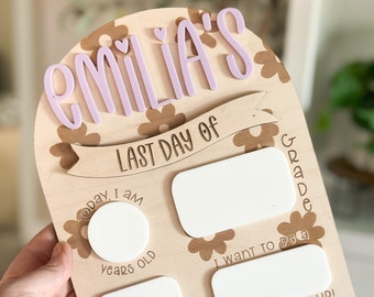 May include: A wooden sign with a pink "EMILIA'S" at the top. The sign has a floral pattern and says "Last Day of Grade" and has blank spaces for the child's age, class, and future aspirations.