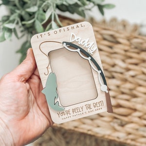 May include: A wooden photo frame with a fishing rod and fish design. The frame has the text "It's ofishal, Daddy You're Reely the Best! Happy Father's Day 2024".