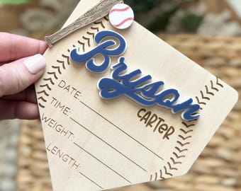 May include: Wooden baseball-themed baby announcement plaque with a blue and white baseball and bat design. The plaque has spaces to fill in the baby's name, date, time, weight, and length. The name "Bryson Carter" is written on the plaque.