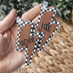 May include: Two black and white checkered lightning bolt shaped keychains with brown leather accents. The keychains are personalized with the names "Jax" and "aiden".