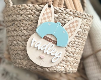 May include: A wooden Easter basket tag shaped like a bunny wearing a blue baseball cap. The tag says "Radley" in white cursive lettering.