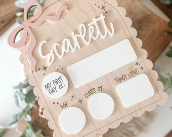 May include: Wooden milestone board with a scalloped edge, a pink bow, and the name "Scarlett" in white. Includes spaces for "My First Day Of", "Age", "Class Of", and "Things I Love".
