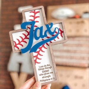 May include: Wooden cross-shaped wall decor with a baseball theme. The cross features a baseball design with red stitching and the name "Jax" in blue. The text "I can do all things through Christ who gives me strength Philippians 4:13" is also included.