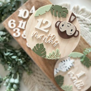 May include: Wooden milestone discs with jungle theme. The 12-month disc features a monkey, green leaves, and the text "12 months". A smaller disc shows a zebra and the text "1 month". Other wooden numbers are visible.