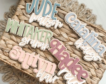 May include: A collection of wooden name tags with various names, including Jude Watson, Whitaker Graham, Autumn Blair, Hadley Reid, and Catalina Indigo. The tags are painted in pastel colors and have a rustic, handmade look.