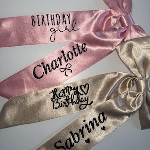 Könnte beinhalten: Drei Satinbänder mit schwarzem Text. Das erste Band ist rosa und sagt "Birthday girl" und "Charlotte". Das zweite Band ist goldfarben und sagt "Happy Birthday" und "Sabrina".