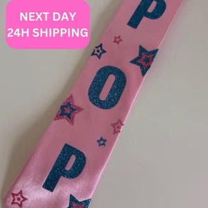 Puede incluir: Una banda de satén rosa claro con la palabra POP en letras grandes de purpurina azul oscuro. La banda está decorada con múltiples estrellas pequeñas de purpurina azul oscuro y rosa, esparcidas por toda la superficie de la tela.